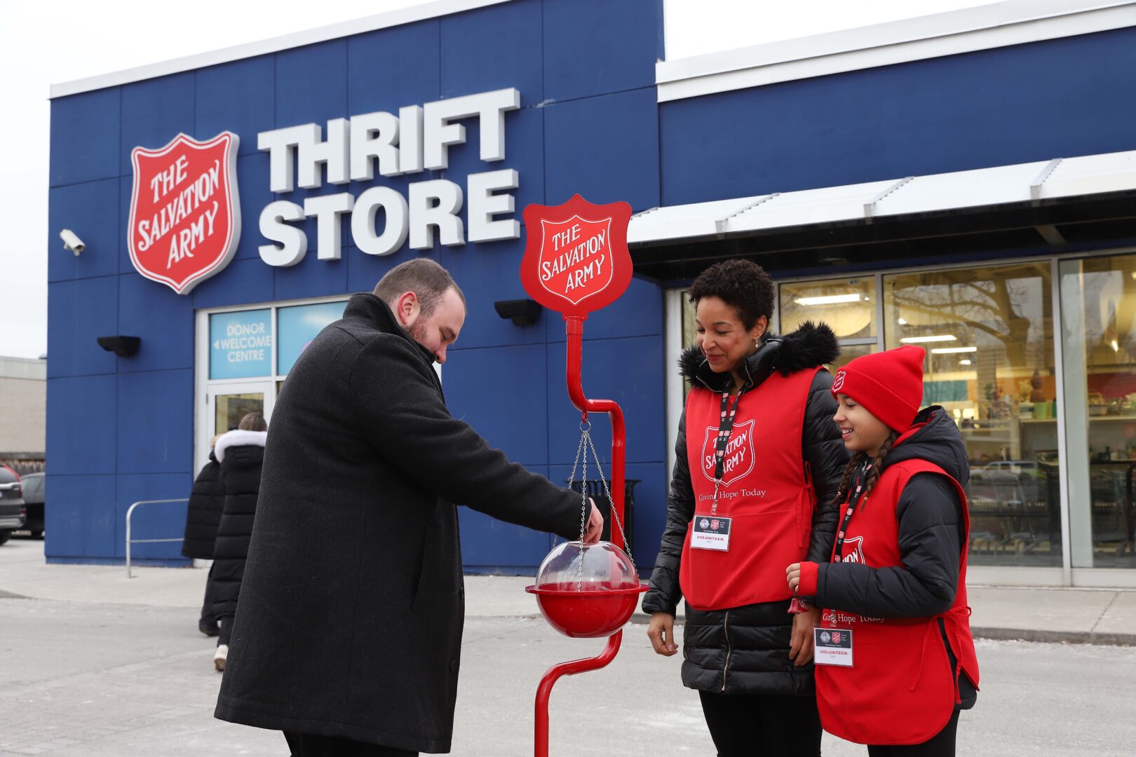 An adult and a youth Salvation Army Christmas kettle volunteer, smiling to donor outside a Salvation Army thrift store.