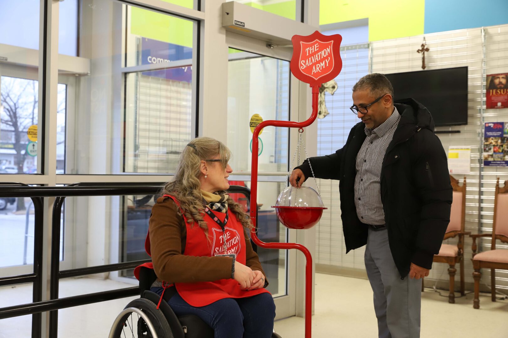 A Salvation Army volunteer in wheelchair smiles at individual donating to kettle inside a shop.